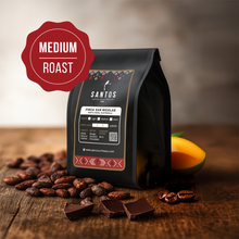 Load image into Gallery viewer, Santos Coffee San Nicolás Medium roast coffee bag, surrounded by cocoa beans, chocolate pieces, and ripe mango slices on a wooden table.