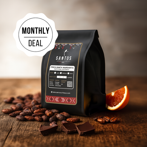 Santos Coffee Monthly Deal showing one bag, surrounded by cocoa beans, chocolate, and an orange slice on a wooden table.