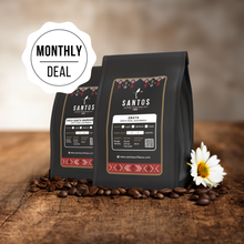 Load image into Gallery viewer, Santos Coffee Monthly Deal showing two bags of coffee, surrounded by roasted coffee beans and a white daisy flower on a rustic table.
