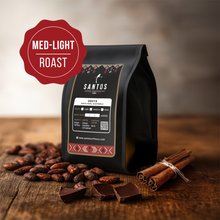 Load image into Gallery viewer, Santos Coffee Obata Medium-Light roast coffee bag, surrounded by cocoa beans, cinnamon sticks, and chocolate pieces on a rustic wooden table.
