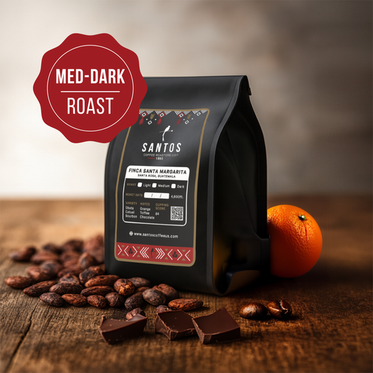 Santos Coffee Santa Margarita Medium-Dark roast coffee bag, surrounded by cocoa beans, chocolate pieces, and an orange on a wooden table.