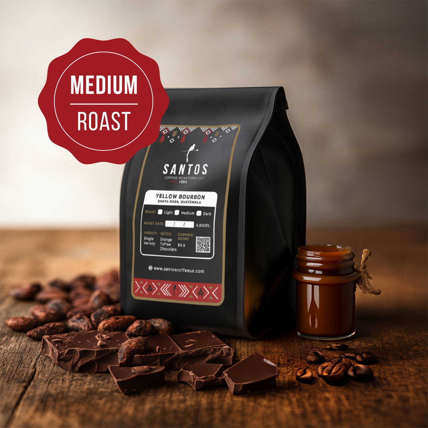 Santos Coffee Yellow Bourbon Medium roast coffee bag, surrounded by cocoa beans, chocolate, and a small jar of caramel on a wooden table.